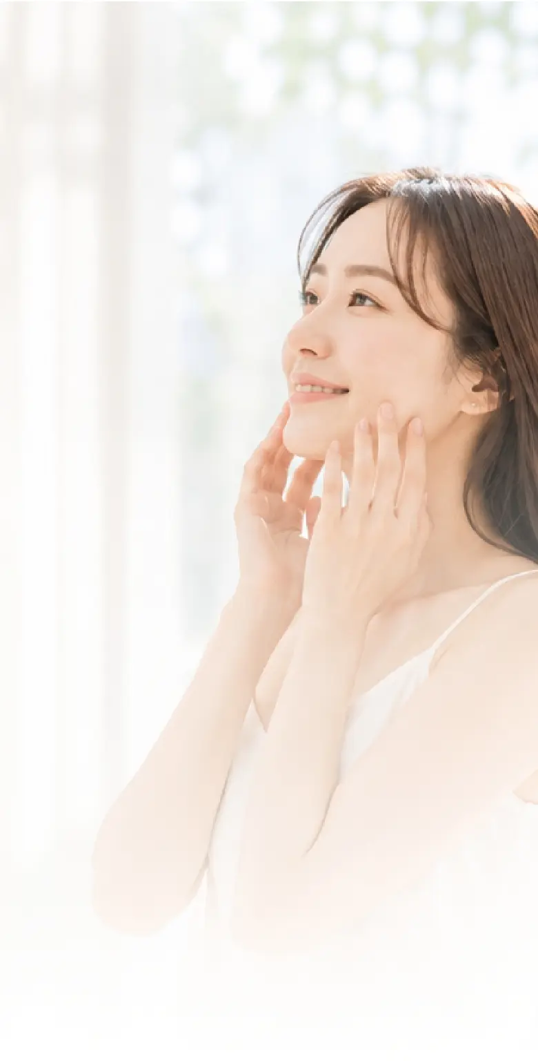 白いキャミソールを着用し、両手で優しく頬に触れながら、明るい表情で横を向く女性